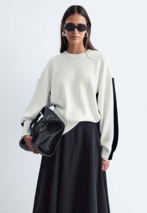 Femme portant des lunettes de soleil noires, un pull blanc avec des rayures noires sur les manches, une jupe noire, tenant un grand sac tressé noir.