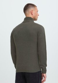 Man gezien vanaf de achterkant met een olijfgroen geribbeld coltrui en zwarte broek tegen een effen lichte achtergrond.