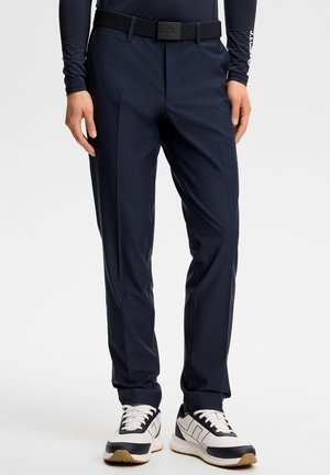 Mann mit marineblauer Hose, schwarzem Gürtel, marineblauem Langarmshirt und weißen sowie schwarzen Turnschuhen steht vor neutralem Hintergrund.