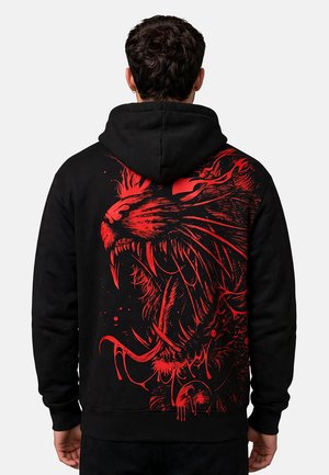 Hombre con sudadera negra con capucha y gran gráfico rojo de tigre rugiente en la espalda, de espaldas a la cámara.