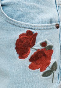 Κόκκινα κεντημένα τριαντάφυλλα με πράσινα φύλλα σε ανοιχτόχρωμο μπλε τζιν ύφασμα κοντά σε τσέπη και πριτσίνι.