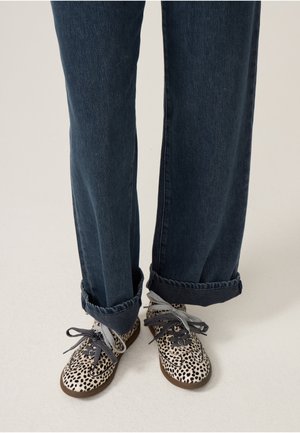 Benen in donkerblauwe opgerolde spijkerbroeken met witte schoenen met zwarte onregelmatige vlekken en dikke zwart-wit gestreepte veters.