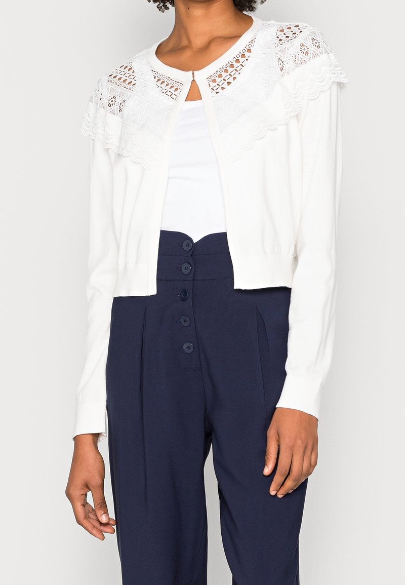 Bílý kardigan s krajkovými detaily na ramenech, zapínání na knoflíky u krku, dlouhé rukávy, doplněný tmavě modrými kalhotami s vysokým pasem.