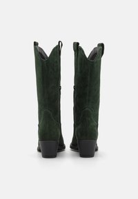 Botas de vaquero verdes de ante con punta afilada, parte superior con ondas y costuras decorativas, que cuentan con un tacón bajo y apilado y un cierre de cremallera.