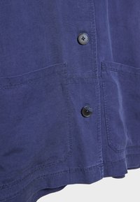Navy blue shirt with two front patch pockets, a rounded hem, and black buttons. Made of lightweight fabric with a smooth texture.