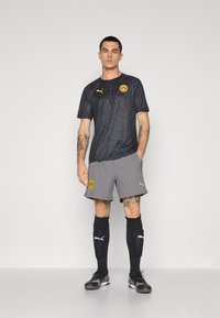 Sort mønstertrøje med gult logo, parret med grå shorts og sorte sokker. Har Puma-branding og et moderne atletisk design.