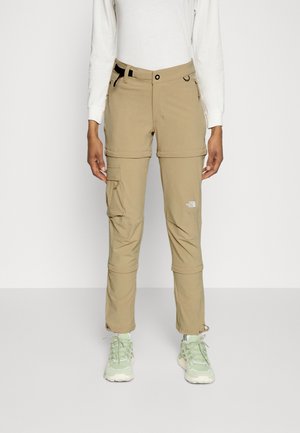 Beige vandrebukser med lynlås ved knæene, cargolomme og logo, båret med lysegrønne sneakers og en hvid bluse med lange ærmer.