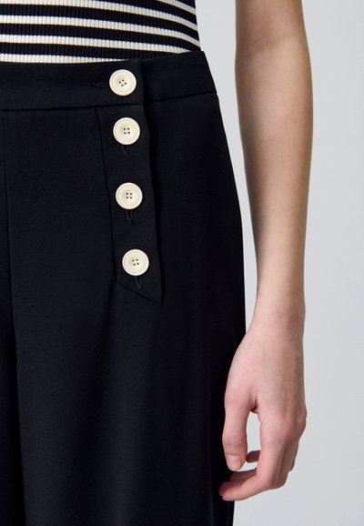 Pantaloni neri con chiusura laterale a bottoni, dotati di quattro grandi bottoni di colore crema. Tessuto morbido con un taglio sartoriale, che crea una silhouette elegante.