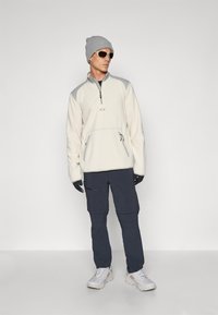 Fleecepullover i lys krem med grå detaljer, med en kvart glidelås. Kombinert med marineblå cargo-bukser og hvite joggesko, accessorert med solbriller og en grå lue.