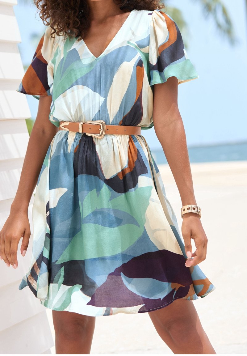 Femme vêtue d'une robe à manches courtes avec un motif de feuilles multicolores, ceinture beige et montre en or, debout à l'extérieur près de l'eau.