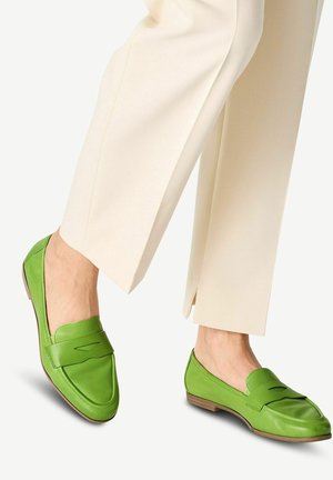 Mocassini in pelle verde con tacco basso e una striscia decorativa sovrapposta, abbinati a pantaloni larghi beige. Texture liscia e design elegante.