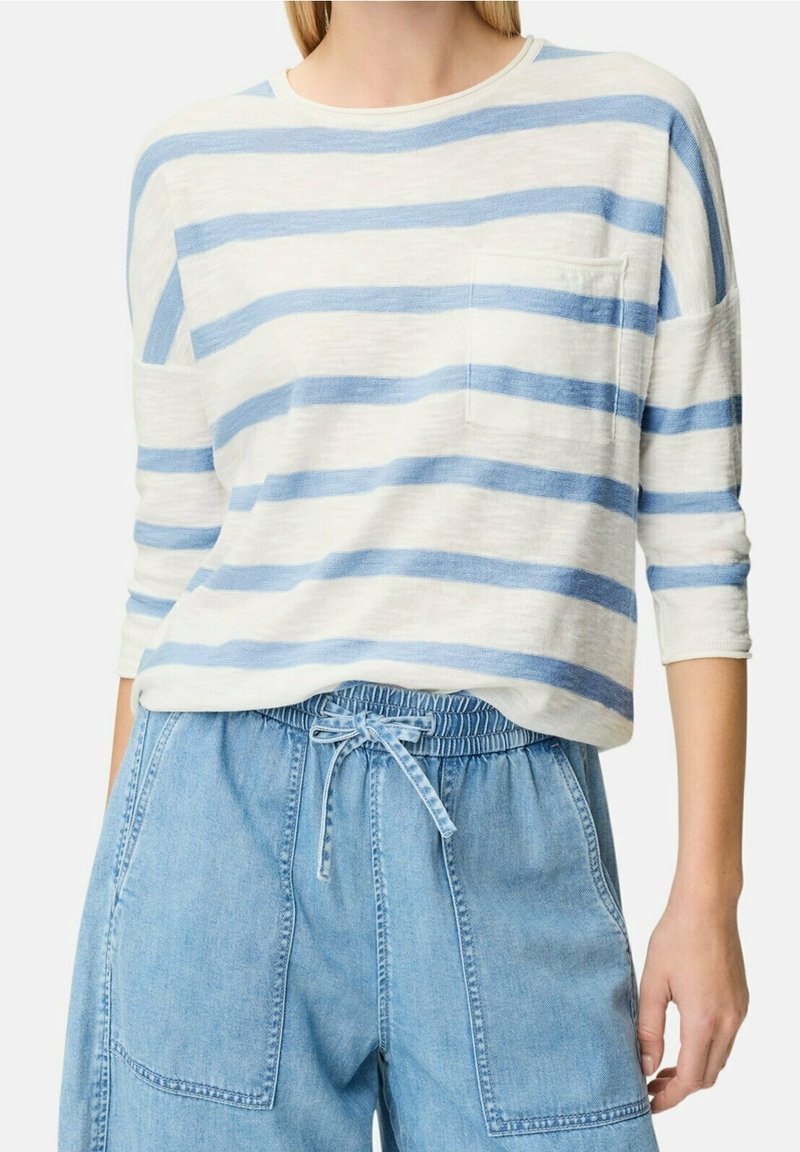 Femme portant une chemise blanche à manches longues avec des rayures horizontales bleues et un pantalon en jean taille haute bleu clair avec une ceinture en tissu.