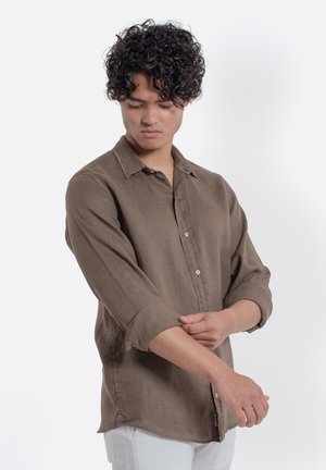 Uomo con capelli ricci che si rimbocca le maniche di una camicia marrone con bottoni su uno sfondo bianco semplice.