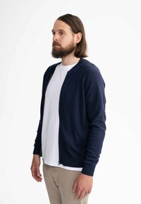 Marineblaue Strickjacke mit Reißverschluss, rundem Ausschnitt und gerippten Bündchen. Getragen über einem weißen T-Shirt mit beigen Hosen.