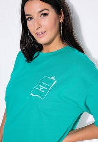 Turkis bomulds t-shirt med et hvidt trykt design af en liste mærket "Kære Santa" på forsiden. Korte ærmer og afslappet pasform.