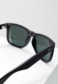 Ochelari de soare din plastic negru, cu lentile gri, cadru pătrat, finisaj lucios, detalii metalice la balama și logo-ul "Ray-Ban" pe bratul ochelarilor.
