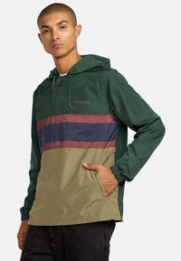 Giacca a vento verde e beige con una striscia orizzontale bordeaux e blu navy, cappuccio con coulisse e tasche laterali.