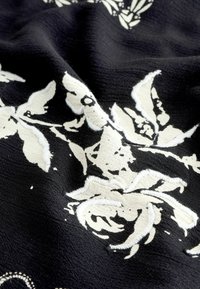 Črn material z belim cvetličnim vzorcem, ki vsebuje vezenine, gladko teksturo in subtilen sijaj. Namenjen za tekstil.