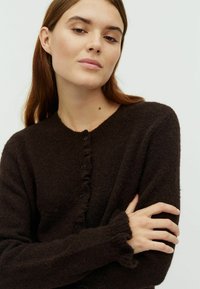 Sötétbarna kötött kardigán kerek nyakkivágással, gombos záródással és fodros szélekkel a mandzsettán és a gomboláson. Puha, texturált gyapjúanyag.