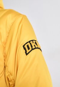 Geltonas nailonas striukė su tekstūruotu juodu siuvinėtu pleistru ant peties su užrašu "DKNY." Pasižymi lygiu audiniu ir siūlėmis.