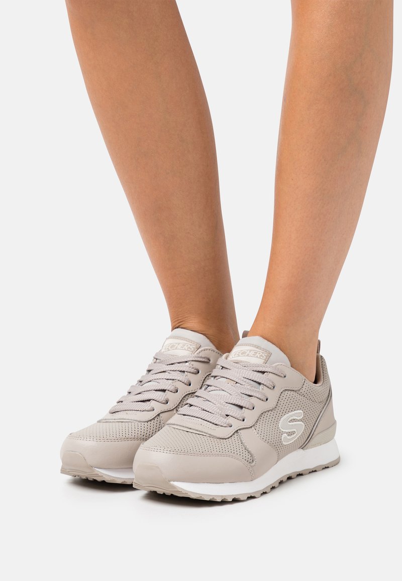 Pies que llevan zapatillas Skechers de color beige claro, con malla y cuero sintético, de pie sobre una superficie blanca.