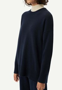 Mörkblå tröja i texturerad stickad kvalitet med ribbat nederkant och rundad halsringning, lagd över en ljusfärgad polotröja.