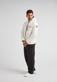Giacca a vento bianca con cappuccio, dotata di chiusura con zip, dettagli neri e un'etichetta di branding rossa. Indossata con pantaloni neri e scarpe bianche.