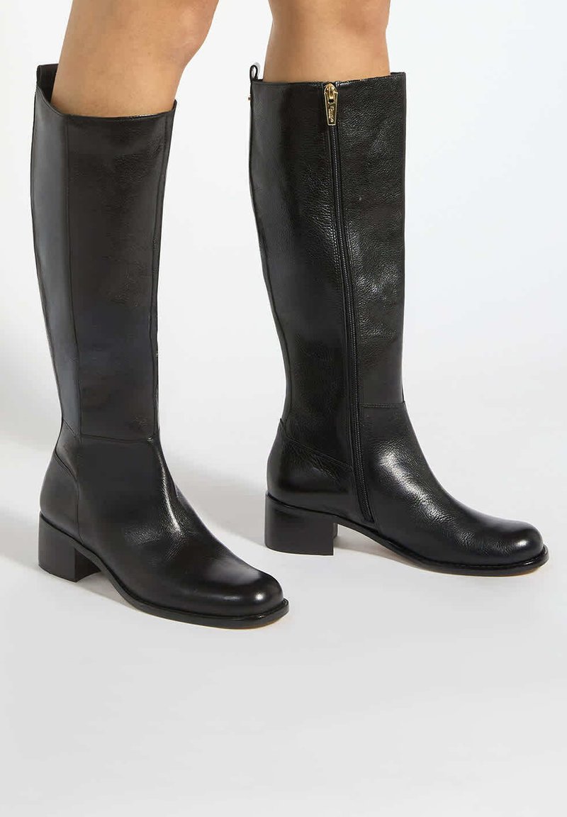 Bottes en cuir noir montant jusqu'aux genoux portées sur les jambes, avec une fermeture éclair latérale et des petits talons blocs sur un fond clair.