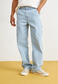 Pantalon en denim bleu clair avec une coupe décontractée, doté de poches avant et d'une texture de lavage subtile. Associé à des baskets beiges.