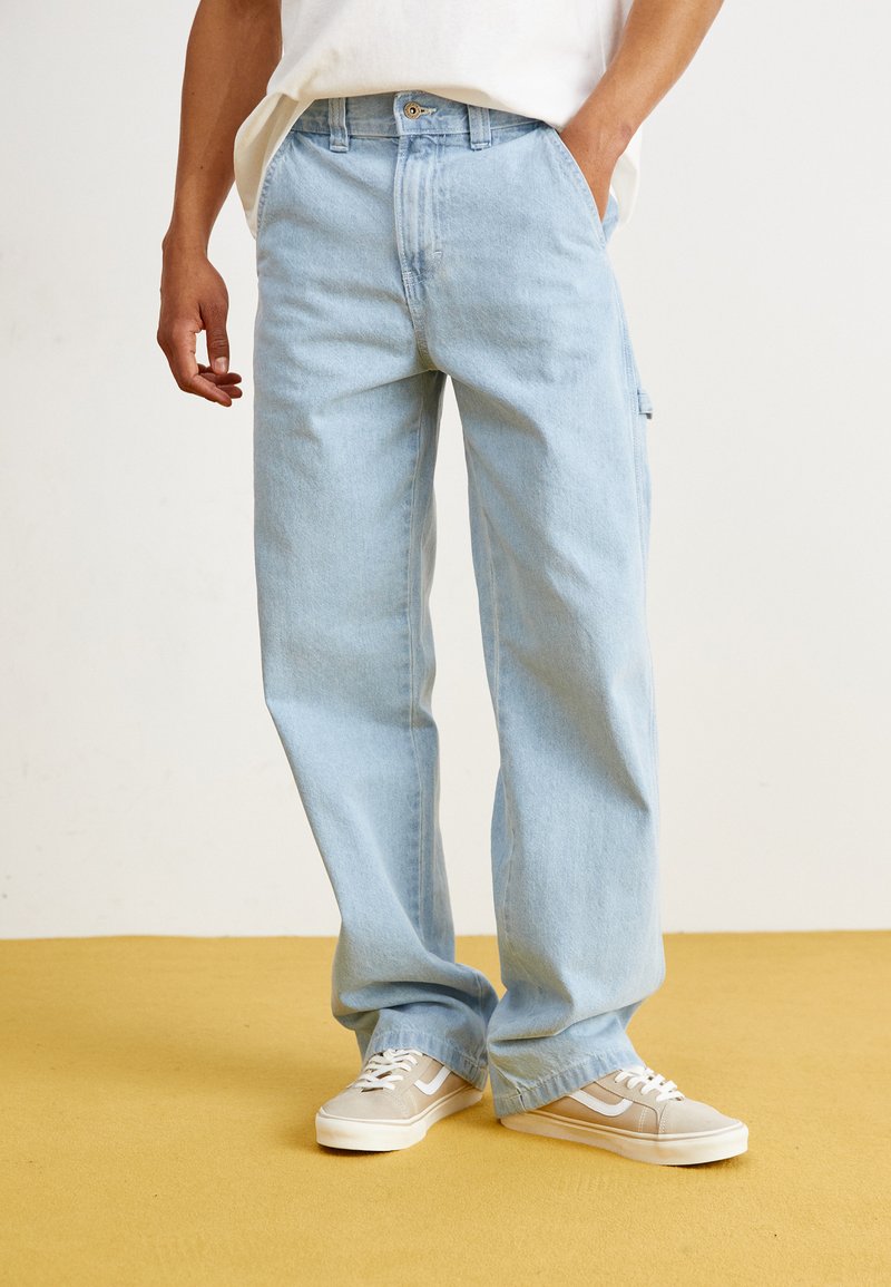 Pantalon en denim bleu clair avec une coupe décontractée, doté de poches avant et d'une texture de lavage subtile. Associé à des baskets beiges.