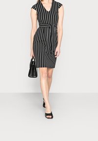 Černobílé pruhované šaty s výstřihem do V, s krátkými rukávy, páskem v pase a sukní sahající do poloviny lýtek, doplněné malou černou kabelkou a otevřenými sandály.