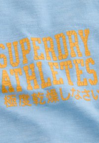 Heledad sinine puuvillakangas, millel on silmatorkav pragunenud kollane tekst "SUPERDRY ATHLETES" koos Jaapani märkidega all, luues tekstuurse visuaali.
