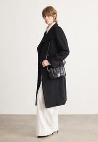 Schwarzer Wollmantel mit strukturiertem Design, breiten Revers, kombiniert mit einer weißen Bluse und einer cremefarbenen, weit geschnittenen Hose. Trägt eine kleine schwarze Handtasche.