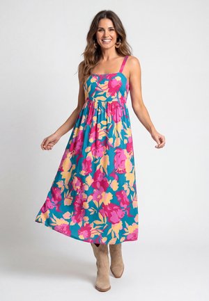 Lächelnde Frau trägt ärmelloses blaues Midikleid mit rosa- und gelbem Blumenmuster und beige Knöchelstiefel vor weißem Hintergrund.