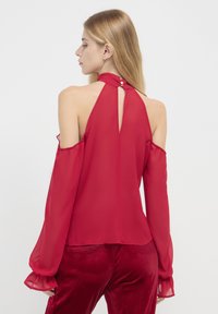 Rote, durchsichtige Bluse mit Schulterfreien Design, langen Ärmeln und einer Knopfschließe am Nacken. Glatte Textur und fließende Silhouette.