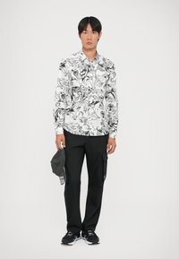 Camicia bianca con motivi neri a spirale, pantaloni cargo neri e sneakers nere. Tiene un cappellino grigio con del testo. Design semplice e casual.