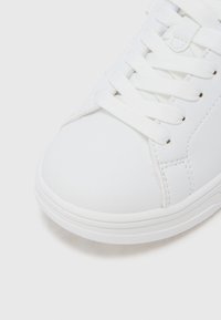 Κοντινό πλάνο στη μύτη ενός λευκού αθλητικού παπουτσιού με υφή στη λαβή από καουτσούκ και λευκά κορδόνια σε λευκό φόντο.