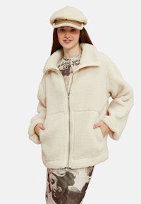 Chaqueta de forro polar blanca con cremallera en la parte delantera, dos bolsillos laterales y cuello alto. Llevada sobre una camiseta gráfica y una falda estampada.
