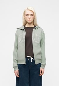 Femme aux cheveux blonds portant un sweat à capuche zippé GAP vert clair, un t-shirt foncé et un jean bleu, debout devant un fond blanc uni.