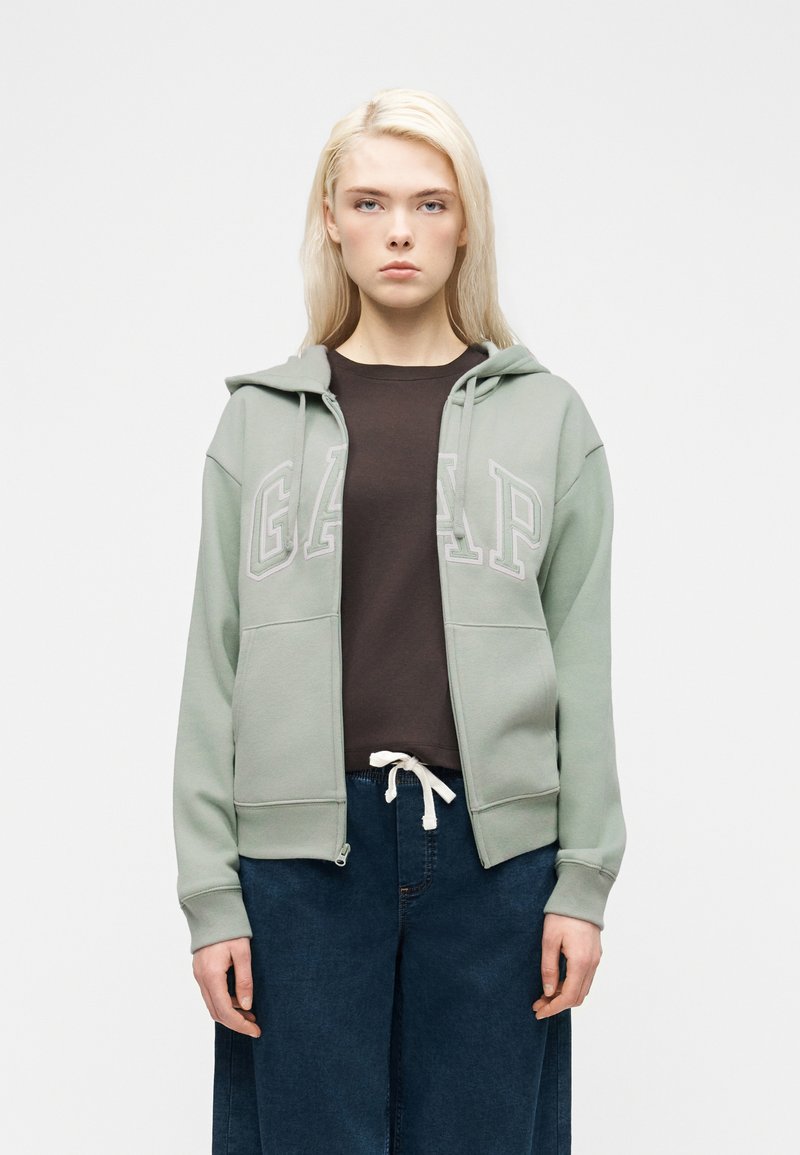 Femme aux cheveux blonds portant un sweat à capuche zippé GAP vert clair, un t-shirt foncé et un jean bleu, debout devant un fond blanc uni.