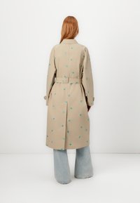Beige trenchcoat met turquoise bloemenborduursel, met een ceintuur in de taille en lange mouwen. Draag het over wijde spijkerbroeken. Aanzicht van achter.