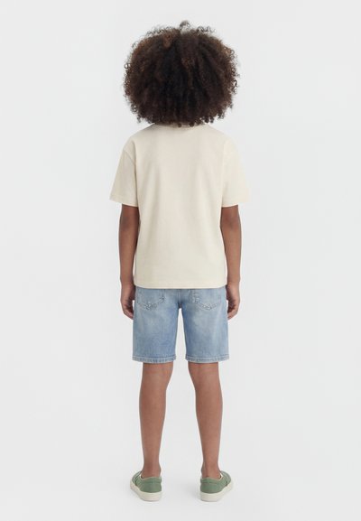 Child with curly hair stands facing away, wearing a beige t-shirt, light denim shorts, and green slip-on shoes.