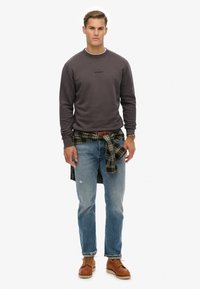 Szary sweatshirt z okrągłym dekoltem, niebieskie dżinsy i brązowe buty. Koszula w kratę związana w talii, prezentująca swobodny, warstwowy look.