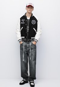 Jeune homme portant une veste de varsity noire et blanche, un jean gris avec de longs cordons blancs, des mocassins noir et blanc, et une casquette de baseball rouge-blanc-bleu.