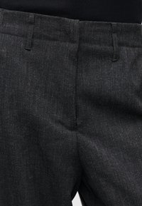 Κοντινό πλάνο της περιοχής μέσης σκούρων γκρι υφασμάτινων παντελονιών με θηλιές για ζώνη και κρυφό εμπρόσθιο κούμπωμα πάνω σε μαύρο φόντο.