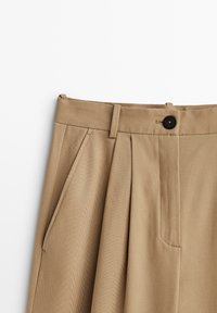 Beige plisserade byxor i strukturerad tyg. Har en midja med en svart knapp och sidfickor.