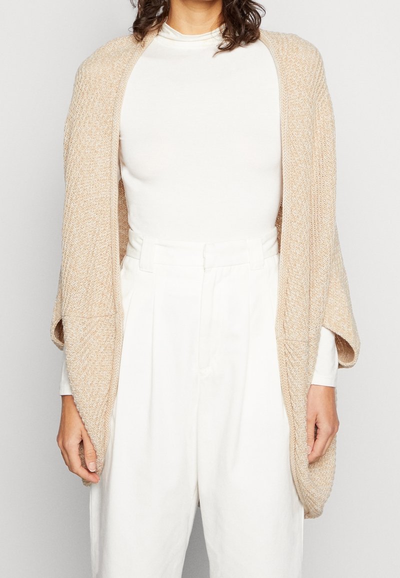 Femme portant un cardigan beige en tricot à ouverture devant, par-dessus un haut blanc à manches longues et un pantalon blanc taille haute, debout devant un fond uni.