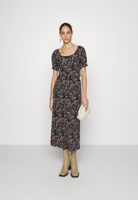Vestido maxi preto com estampado floral e mangas bufantes, apresentando padrões em rosa e verde, cintura ajustada e fecho de botão na frente. Usado com botas bejes até ao joelho.