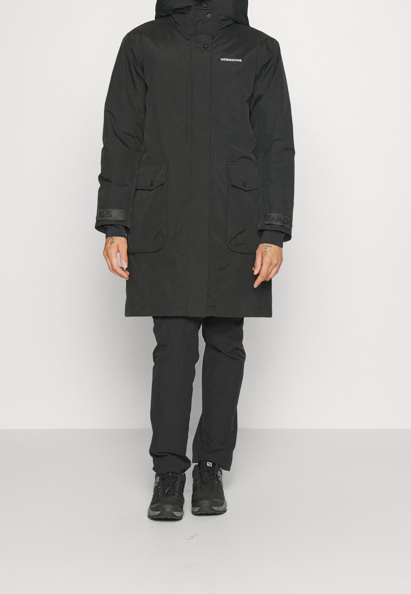 Personne portant un manteau noir longueur genou avec poches à rabat, pantalon noir et chaussures de randonnée noires, debout devant un fond blanc.