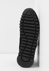 Semelle de chaussure noire avec un motif de bande de roulement épais et rainuré conçu pour la traction, présentée verticalement sur un fond d’angle blanc.